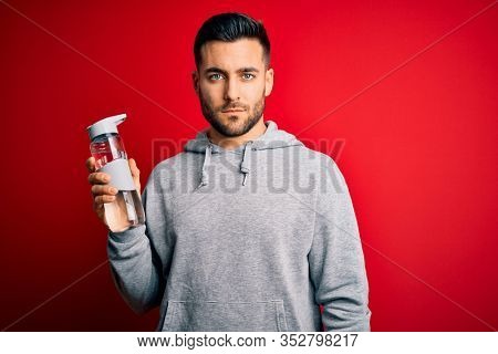 Young handsome sportsman drinking bottle of water to refeshment over red background with a confident expression on smart face thinking serious