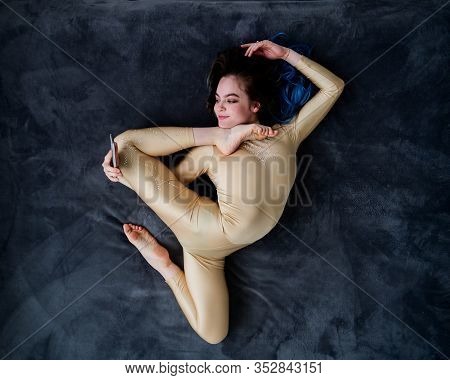 A Female Acrobat Lies On A Gray Bedspread In A Curved Pose And Uses A Mobile. A Woman Contortionist 
