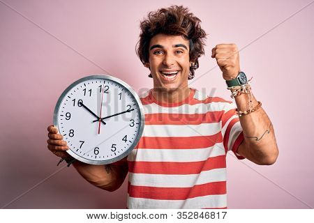 Young handsome man holding big clock standing over isolated pink background screaming proud and celebrating victory and success very excited, cheering emotion
