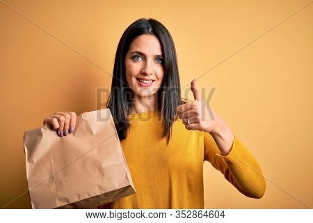 Young brunette woman with blue eyes holding delivery paper bag with food happy with big smile doing ok sign, thumb up with fingers, excellent sign