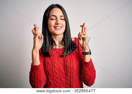 Young brunette woman with blue eyes wearing casual sweater over isolated white background gesturing finger crossed smiling with hope and eyes closed. Luck and superstitious concept.