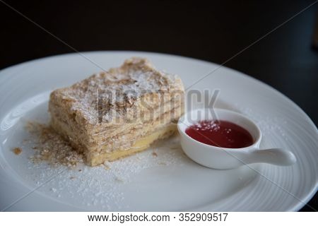 A Piece Of Napoleon Cake On A White Plate. The Cake Is Sprinkled With Powdered Sugar. There Is A Cup