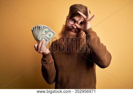 Redhead Irish man with beard holding a bunch of 1 dolla banknotes over yellow background with happy face smiling doing ok sign with hand on eye looking through fingers