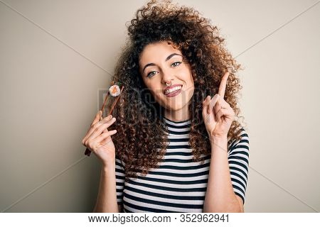 Young beautiful woman with curly hair and piercing eating sushi using wooden chopsticks surprised with an idea or question pointing finger with happy face, number one
