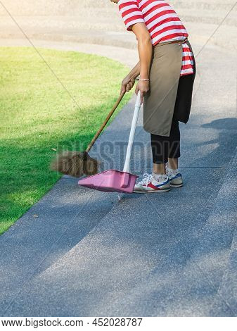 Female Worker Sweeping Yard With Broom Tool And Dustpan In Ornamental Backyard Garden. Woman Wear Ap