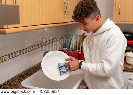 Caucasian Young Man With Curly Hair Washing The Dishes In The Kitchen, His Inexperience In Housework