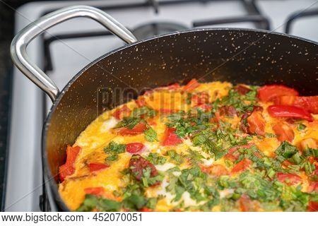 Fried Egg With Vegetables And Herbs In Black Frying Pan On White Stove. Side View