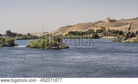 There Are Small Islands In The Riverbed. There Is Green Vegetation On The Shore. In The Distance, Th
