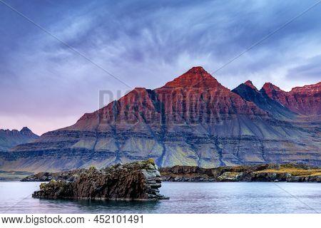 The Bulandstindur, or Pyramid mountain, in the East Fjord region of Iceland. For a few moments, the rock gows red as the sun rises.  A lion shaped rock sits in the water. 