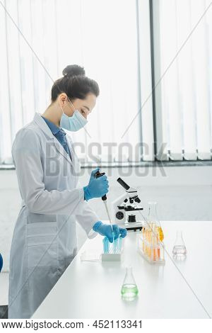 Side View Of Scientist In Medical Mask Working With Micropipette And Test Tubes Near Microscope.