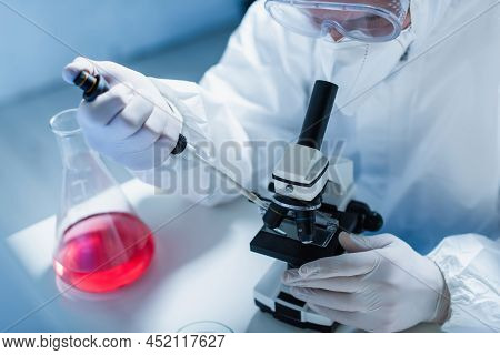 Bioengineer In Hazmat Suit Holding Micropipette Near Microscope And Flask With Liquid.