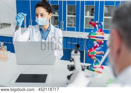 Geneticist In Medical Mask Working With Test Tube And Laptop Near Blurred Colleague.