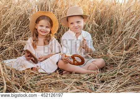 Happy Child Boy And Girl In Straw Hats Sitting In Wheat Field, Eating Bread On Summer. Cottage Core
