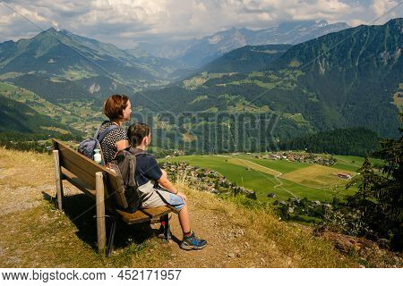 Mother And Son Sitting On A Bench Enjoying Amazing View High In The Vaud Alps While Hiking On A Sunn