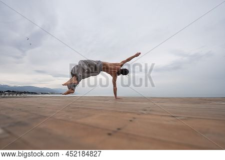 Shirtless Caucasian Man Doing Backflip On Pebble Beach.