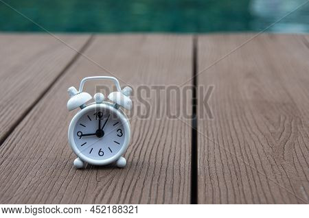 White Alarm Clock On Wooden Floor With Blurred Swimming Pool Background. The Clock Set At 9 O\'clock