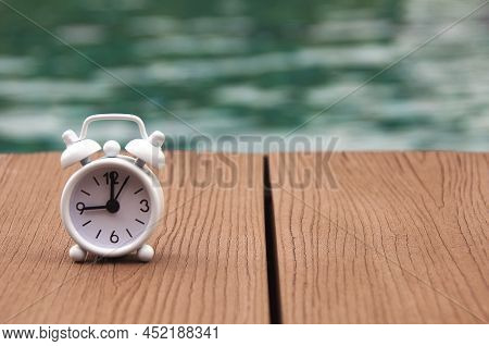 Alarm Clock On Wooden Floor With Blurred Swimming Pool Background. The Clock Set At 9 O\'clock. Morn