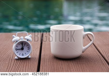 White Alarm Clock On Wooden Floor With Coffee Cup. With Blurred Swimming Pool Background. Clock Set 