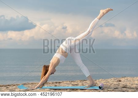 Young Fit Woman In White Sportswear Practices Yoga Asana Pose On The Beach On Bright Sunny Day. Down