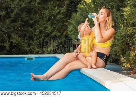 Summer Heat. Young Mother Drinking Water From A Bottle And Holding Her Lettle Daughter. Family Sitti