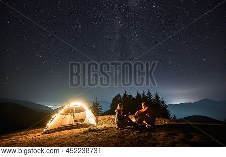 Woman With Guitar In Her Hands Sitting Opposite Man By Campfire Near Their Light Shining Tent. Roman