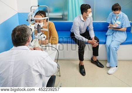 Medical Nurse Talking To Patient When His Wife Getting Eyes Examined With Autorefractometer