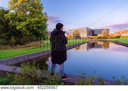 A Young Black-haired Woman In Warm Clothes Stands In A Park On The Edge Of A Water Channel With A Re
