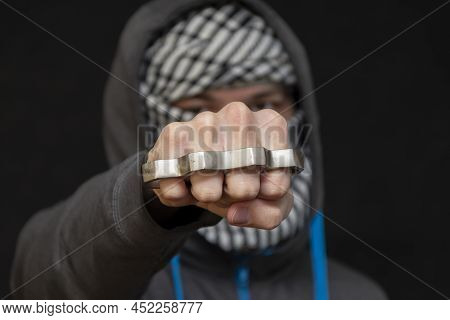 Brass Knuckles In The Hand Of A Man With A Scarf Covering His Face On A Dark Background, Selective F