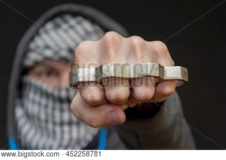 Brass Knuckles In The Hand Of A Man With A Scarf Covering His Face On A Dark Background, Selective F