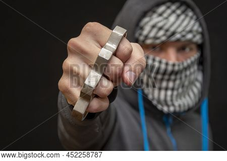 Brass Knuckles In The Hand Of A Man With A Scarf Covering His Face On A Dark Background, Selective F