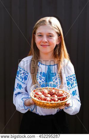 Defocus Ukrainian Smiling Girl In Vyshyvanka Holding Basket Of Colored Red Eggs On Black Background.