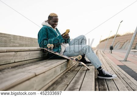 Happy Young African Man With Skateboard Looking At Mobile Phone Social Media App To Connect With Fri