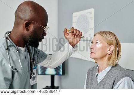 Young confident male opthalmologist in uniform examining retina of mature female patient with astigmatism with help of medical device
