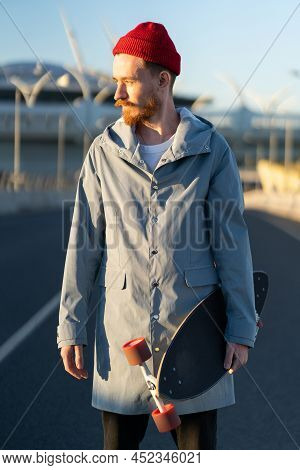 Casual Man With Longboard On Sunset Over Modern City Building. Hipster Skater Holding Skateboard Wea