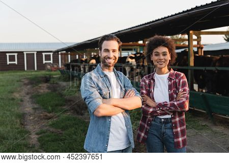 Happy Multiethnic Farmers Standing With Crossed Arms Near Blurred Cowhouse