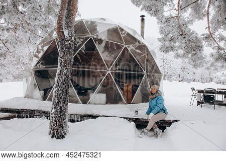 A Woman Sits And Enjoys The View Next To A Dome Tent In Glamping In The Winter Forest.