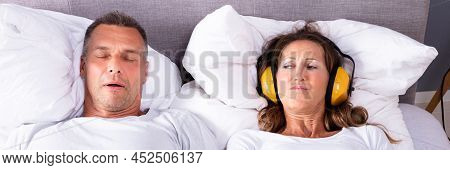 Mature Woman Covering Her Ears With Headphone While Man Snoring In Bed