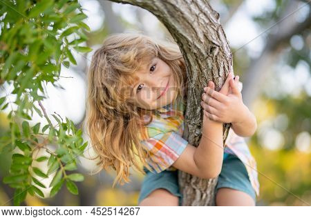 Happy Child Playing In The Garden Climbing On The Tree. Boy Climbs Up The Tree In Summer Park.