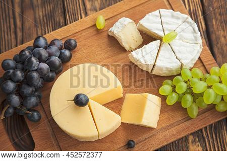 Assorted Cheeses, Camembert And Gouda Cheese On A Wooden Cutting Board With Different Types Of Grape
