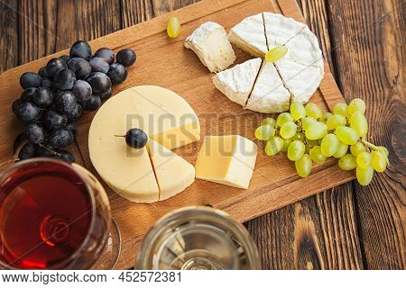 Various Types Of Cheese With Dark And Green Grapes And Wine On Wooden Table.