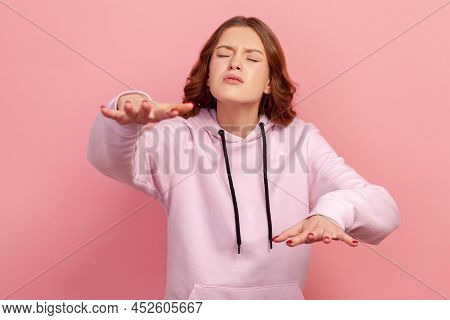 Portrait Of Concentrated Brunette Young Female In Hoodie Trying To Move With Closed Eyes And Raised 