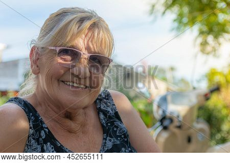 Smiling Blonde Woman On A Sunny Day, Green Trees, Motorcycle In The Background