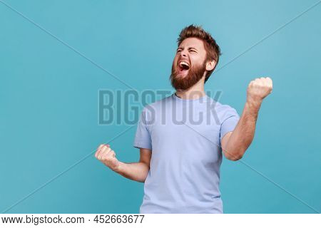 Portrait Of Overjoyed Bearded Man Standing With Excited Expression, Raising Fists, Screaming, Shouti