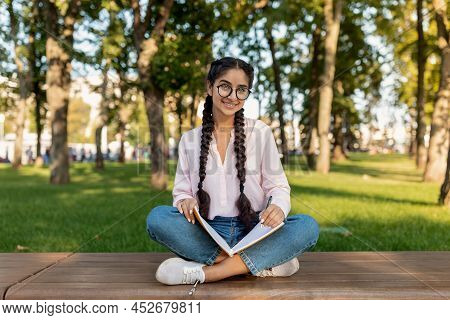 Studentship Concept. Happy Indian Student Girl Doing Homework, Holding Workbook While Sitting On Ben