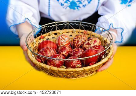 Defocus Female Hands Holding Basket Of Colored Red Eggs On Nature Green Grass Background. Easter Hol