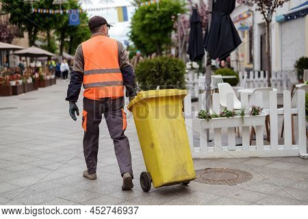 Street Worker Janitor In Orange Uniform With Yellow Garbage Bin.