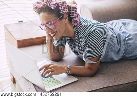 Happy Woman At Home Using Laptop Computer Laying On The Sofa With Pink Curlers On Hair. Concept Of M