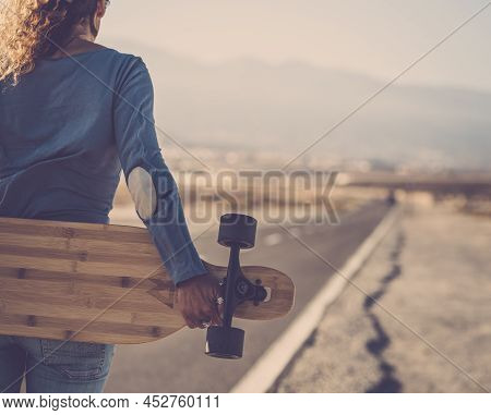 Carefree Woman Standing And Holding Wooden Longboard Behind Her Back On Side Of Long Asphalt Road. A