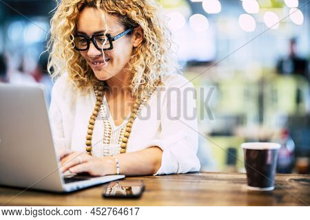 Happy Woman Working On Laptop At Coffee Shop. Woman In Eyeglasses And Beads Necklace Using Laptop On