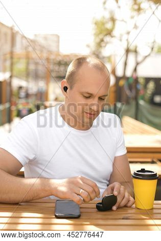 Man Is Sitting At A Table In The Patio Of A Restaurant And Takes Out Wireless Headphones From A Case